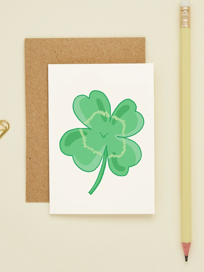 Mini greeting card featuring a hand-drawn four-leaf clover with a smiling face, styled with a kraft envelope and yellow pencil on a cream background.