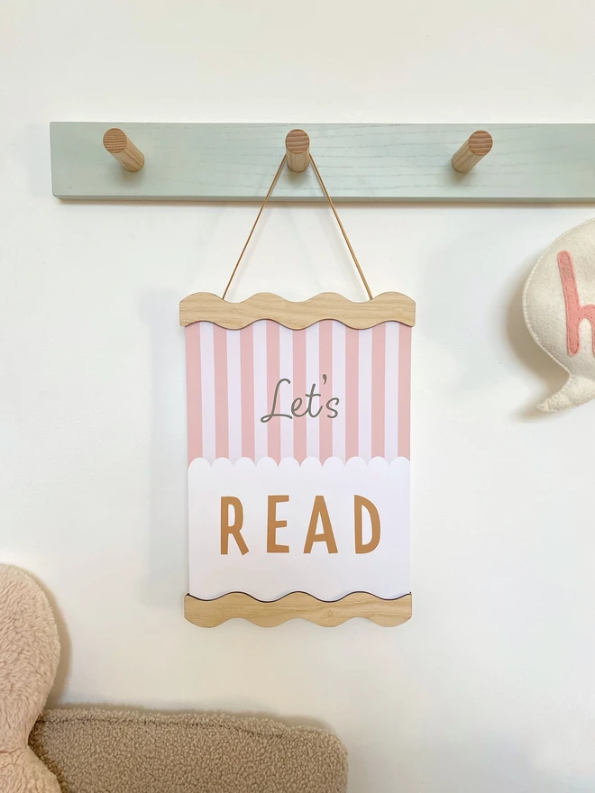 a wooden picture frame hung on a peg rail. the picture frame is wavy wood.  the frame is strung with vegan suede and holds children's colourful print reading Lets Read