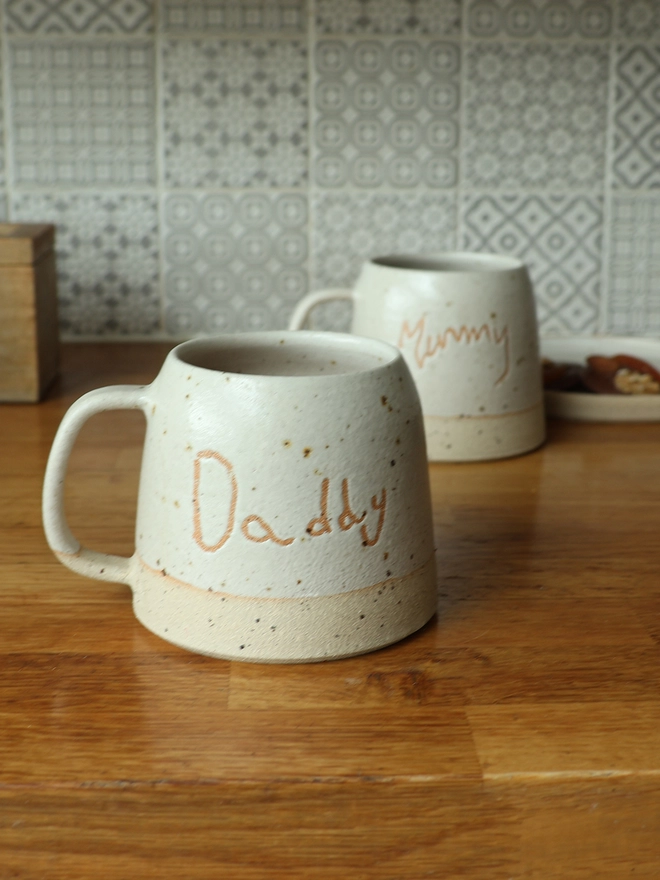 Personalised Kids' Handwriting Handmade Stoneware Mug with the name 'Daddy' on a wooden kitchen counter. The background shows the same mug with the name 'Mummy'.
