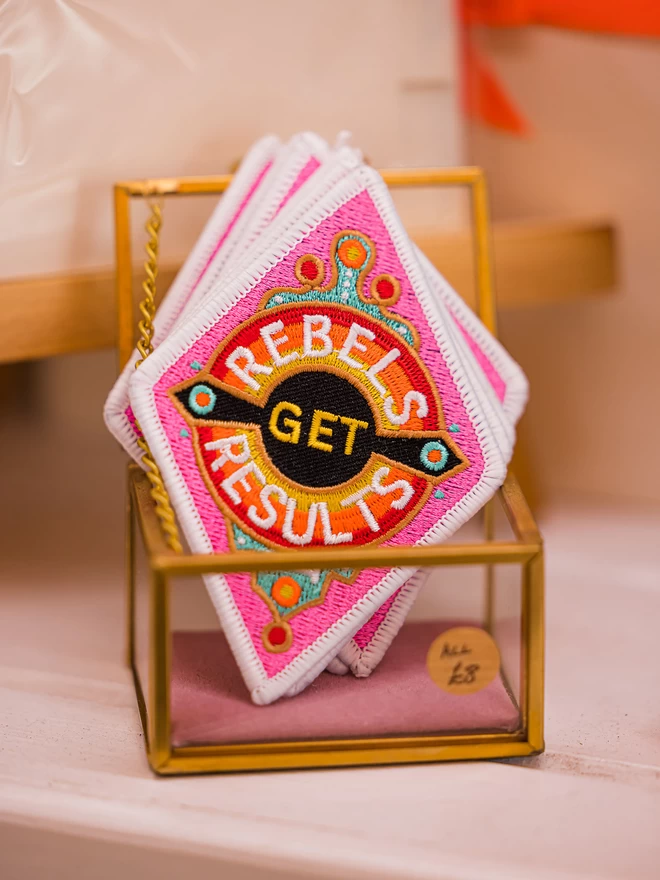 Several patches sit in a gold and see through ornamental box. The patch is a diamond shape embroidered patch with “Rebels Get Results” at the centre. The design features pink, white, red, yellow and black colours and has a white border.