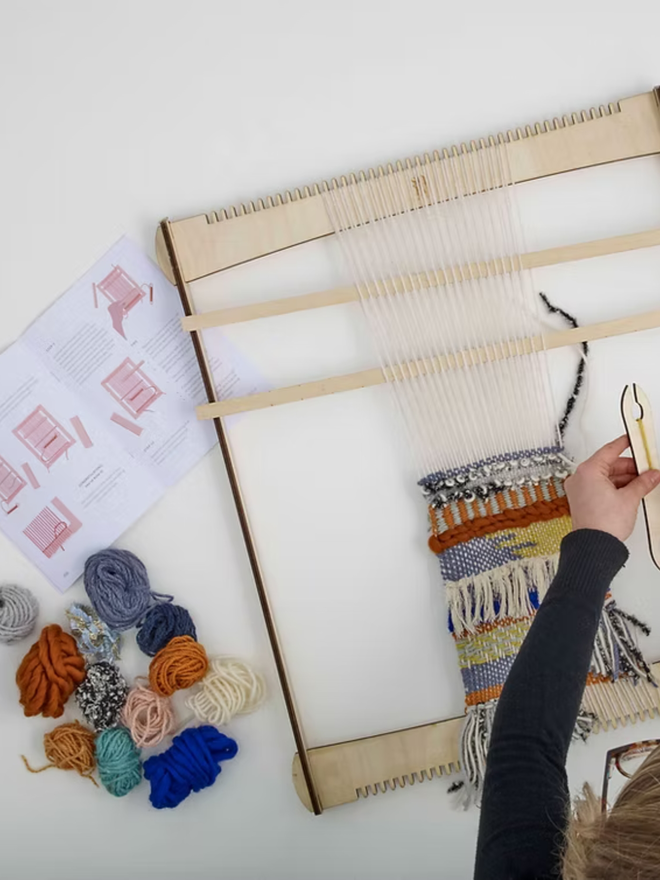 large weaving kit. a large loom on a white surface, with yarn in various colours and a person in the early stages of the weaving process.