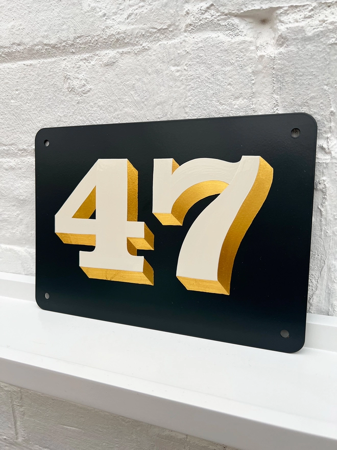 Hand painted house number against a white brick wall, number 47 in serif style in off white and directionally burnished 23 carat gold leaf, on anthracite grey background. 