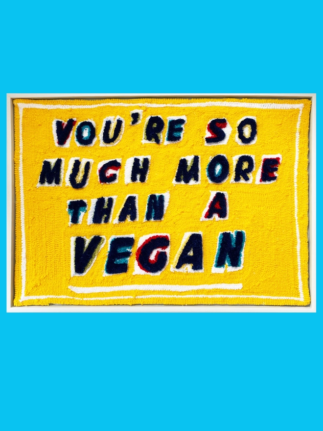 You're so much more than a vegan written in blue tufted lettering with red, blue and yellow tufted shadows on a bright yellow background.  Piece is framed in a white wood frame and sat on a duck blue background