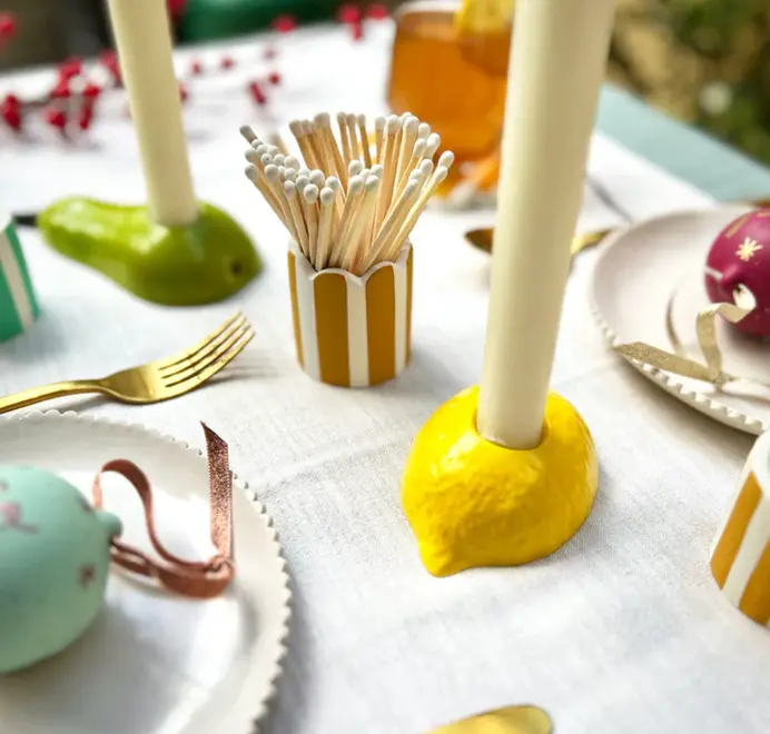Candles placed in yellow lemon candlestick holders displayed on a table alongside matchstick holder. 