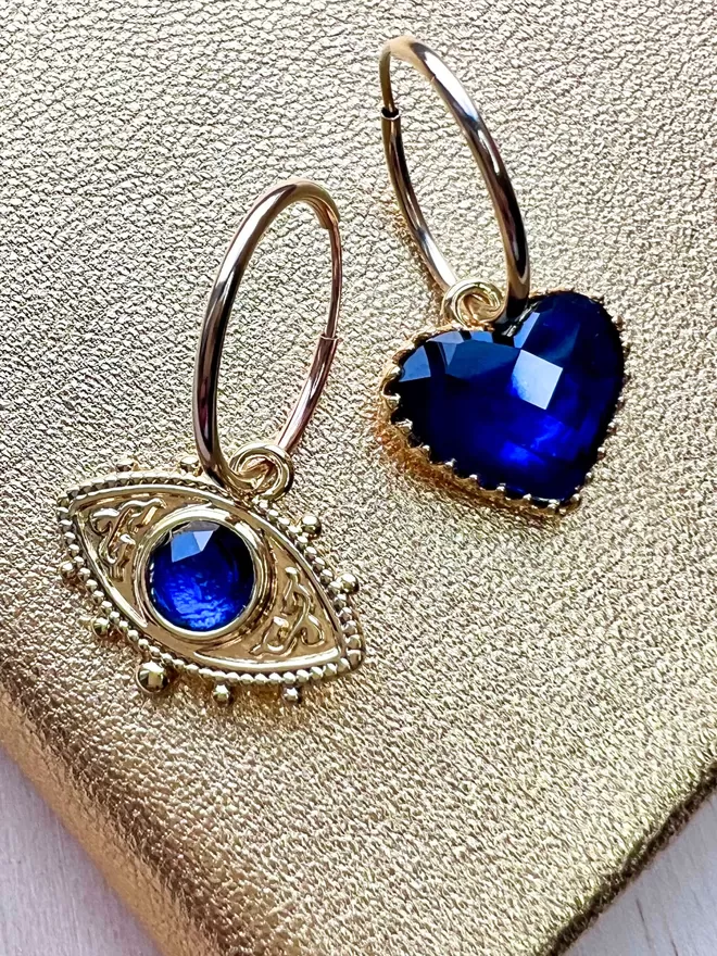 Mismatching gold hoop earrings with blue evil eye charm and blue faceted heart charm on gold backgroundrings 