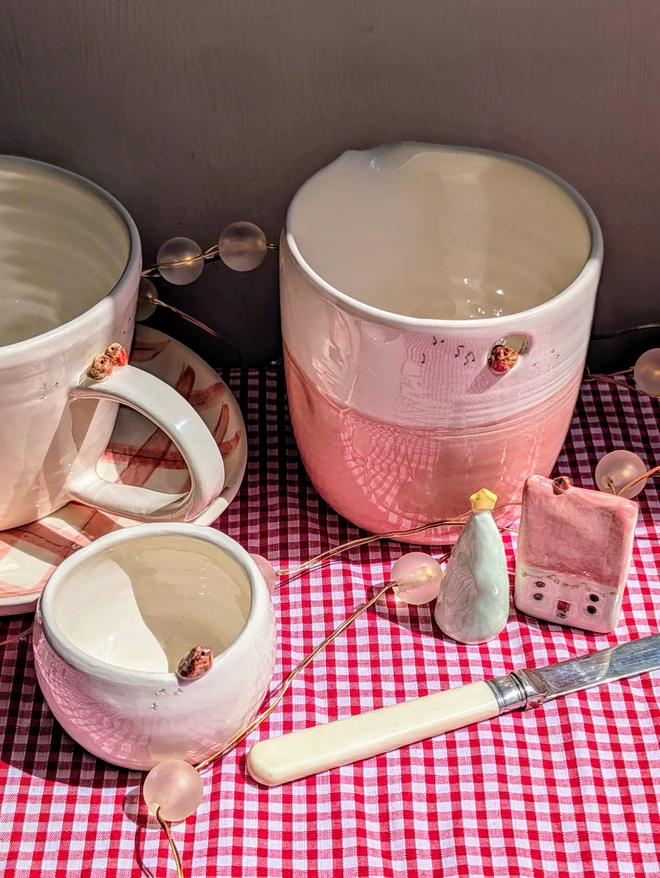 christmas pottery in pink and white with tiny ceramic robin birds and a tiny pottery house and christmas tree ornament all on a red gingham tablecloth