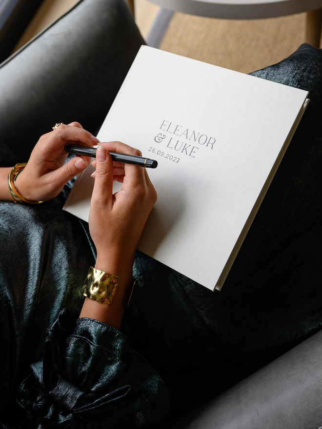 A pearl-coloured wedding guest book, personalised with printed lettering on the cover, rests on a woman's lap.