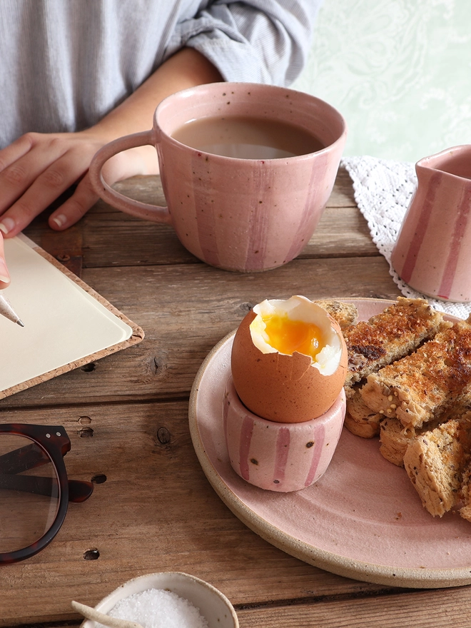 A table scene with a handmade ceramic stoneware mug, milk jug and egg cup, a plate of toast, a pair of glasses and a notebook which a female sits behind with a poised pen. 