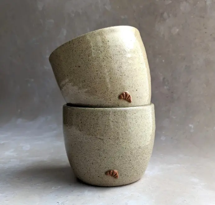 Two croissant coffee cups in biscuit glaze stacked on top of each other against a grey marble background.