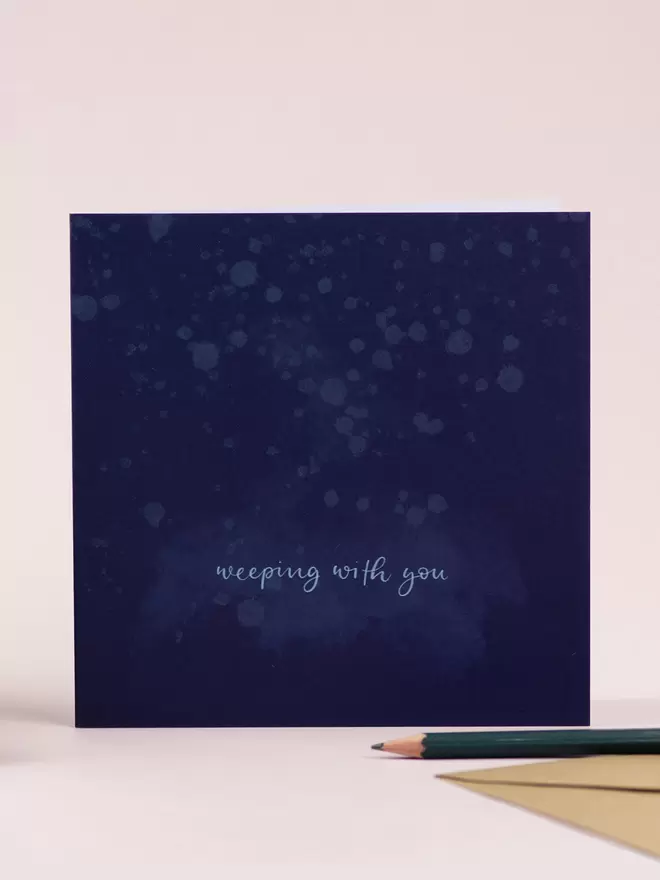 A deep blue card stands against a pale pink backdrop. The card has a subtle water-drop-splatter design and the hand-lettered words 'weeping with you'