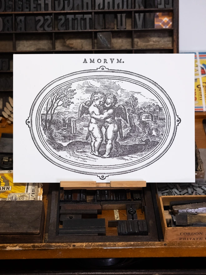 monochrome black illustration of mythological cherubs embracing framed by an ovoid border resembling a wall mirror. Printed above in vintage block font is the word Amorum, styled AMORVM. 