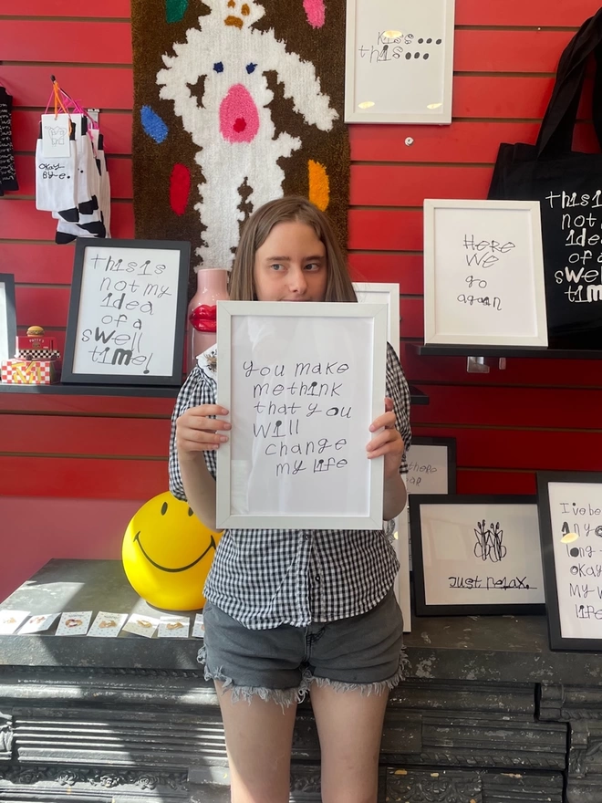 Piper a deaf, autistic teenage artist from hackney holds her artwork in a white frame. her art is black handwritten unique font on a white background.  Piper is stood in front of a lot of her different framed notes in her mums shop.