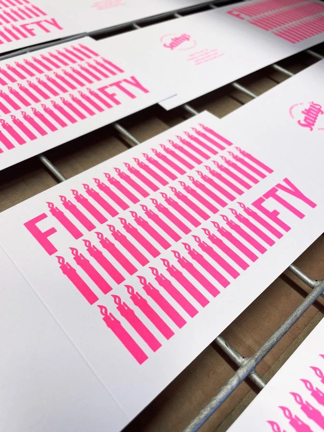 An angled shot of several unfolded screen printed Fifty birthday cards on a drying rack. The word Fifty screen printed in neon pink ink across the rectangular white recycled cards. 