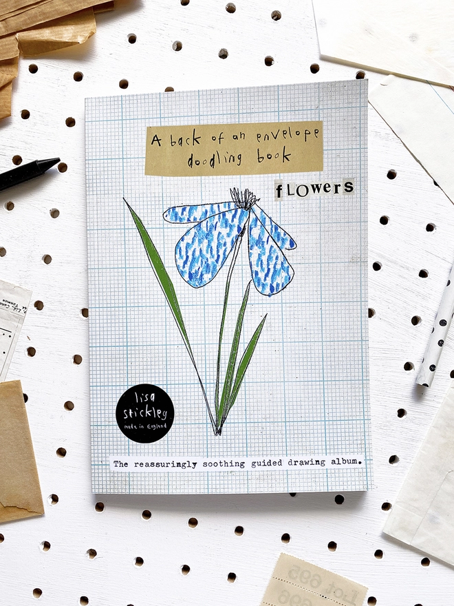 book with blue flower drawing on the cover on white peg board with scattered paper
