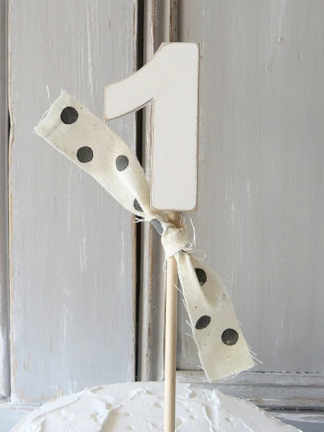hand painted wooden number cake topper. a white number one cake topper with a spotty ribbon on a stack of donuts.