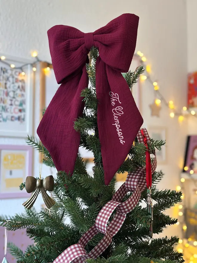 A handmade burgundy Christmas bow tree topper sits at the top of a decorated Christmas tree.
