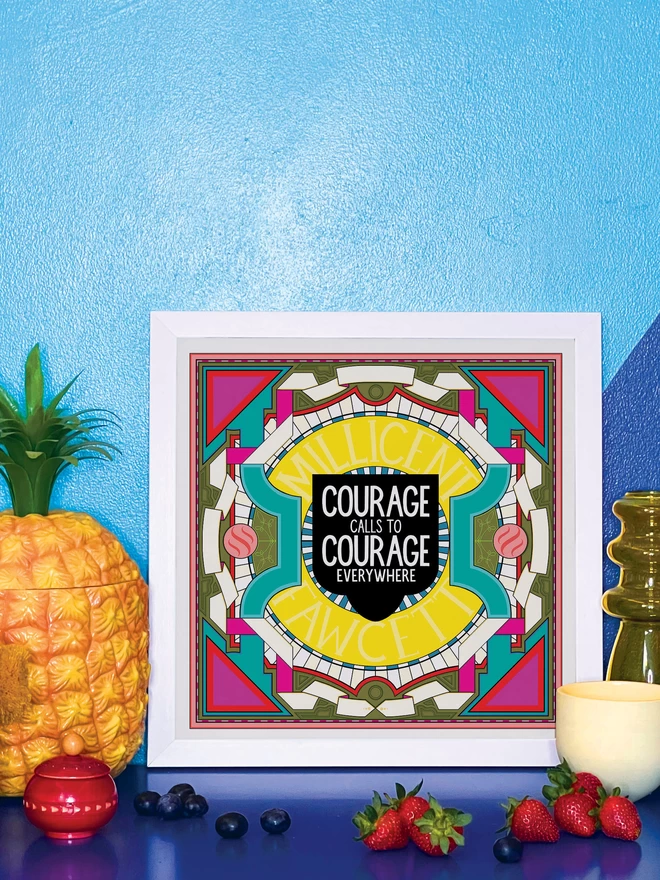 The illustration is in a white square frame with “Courage calls to courage everywhere” written in white against a black background at the centre, surrounded by Millicent Fawcett written in yellow, and bordered with a symmetrical design in white, greens and pinks. It is propped against a wall painted light and dark blue, on a shiny blue cabinet. Next to the frame is a plastic pineapple ice bucket, a small wooden red pot a handful of blueberries, a yellow glass vase, a small yellow ceramic pot and 4 strawberries. 