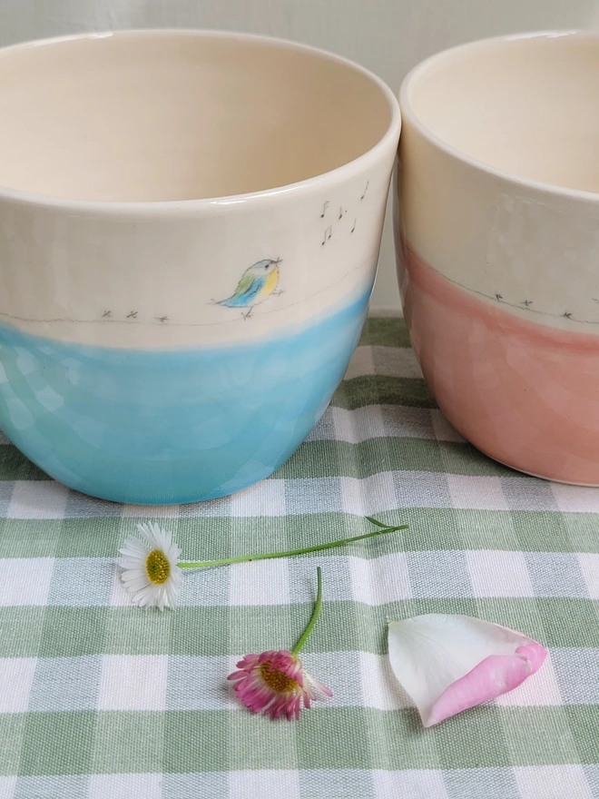 2 pottery beaker cups with blue and pink glaze and hand painted blue tit, musical notes and bird prints on a green gingham table cloth 