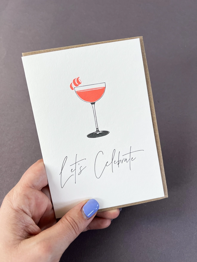 A neon orange Cosmo drink with a cute garnish o the rim of the glass and the words "Let's Celebrate" beautifully written in a modern calligraphy.