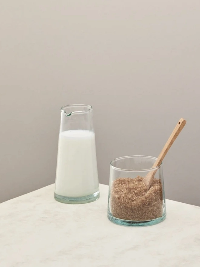 Tall Jug, a tall glass jug sitting on a plain counter top against a white backdrop. It is filled by milk, and accompanied by another smaller container. 