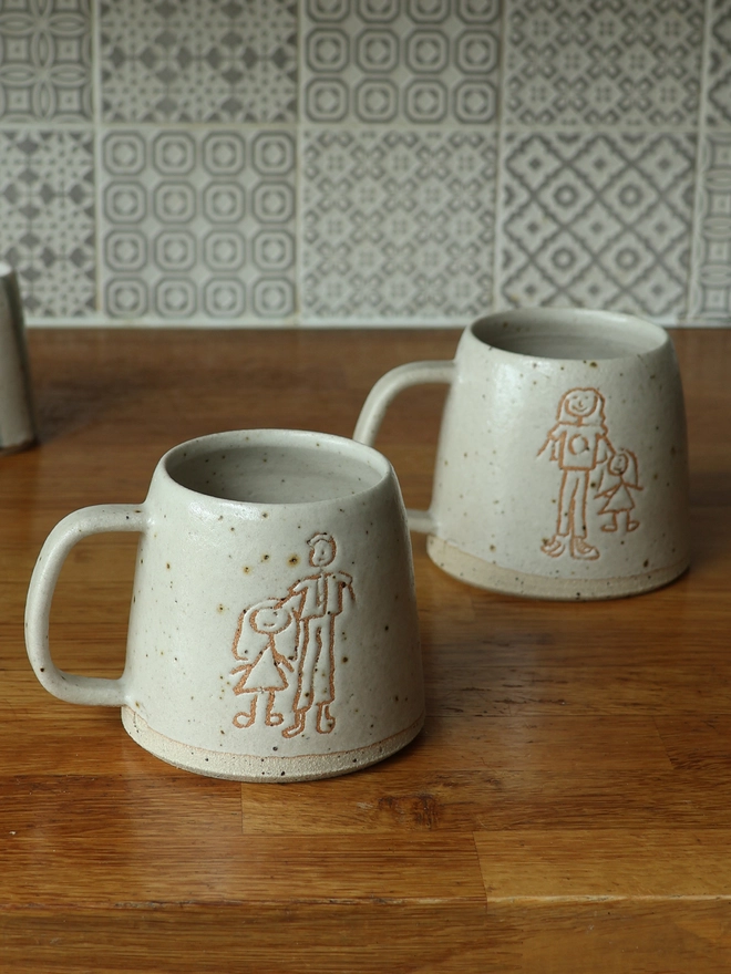 2 Childrens Drawing Handmade Ceramic Mugs on a wooden kitchen surface. The Children's drawings feature parents and children