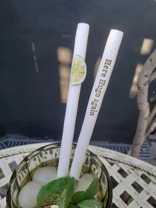 Pair of handmade ceramic cocktail straws in a Hugo Spritz, featuring a lime slice decoration and the phrase “Here Hugo again”, shown in a mint-garnished cocktail.