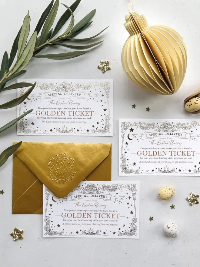 Three Golden Tickets offering a giant Easter egg, a weekend activity and a family movie day, laid out with the gold envelope. Olive leaves, speckled mini eggs and a paper honeycomb decoration surround them.