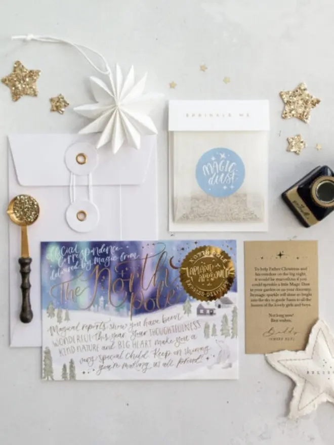 An overhead view of the North Pole notecard with gold foil lettering and a gold seal, a white string-and-washer envelope, a Magic Dust glassine bag with a pale blue label and a kraft instruction card. A paper star, gold glitter stars, a glitter spoon, an ink pot and a felt "Believe" star sit around them.