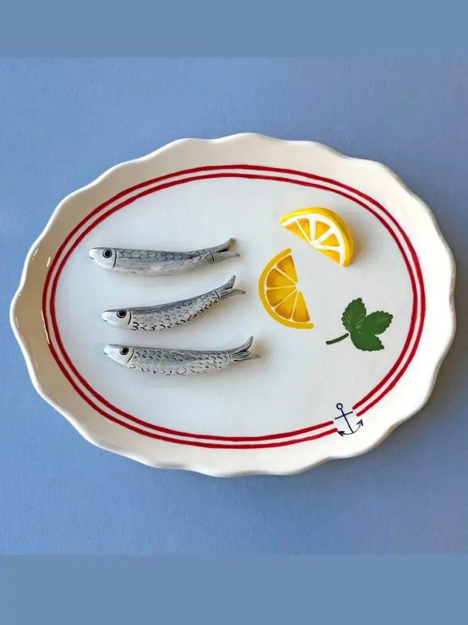 ceramic white platter with red border and anchor detail, with 3 ceramic sardines arranged on top, a ceramic wedge of lemon plus lemon painted on, with parsley detail, on a blue background