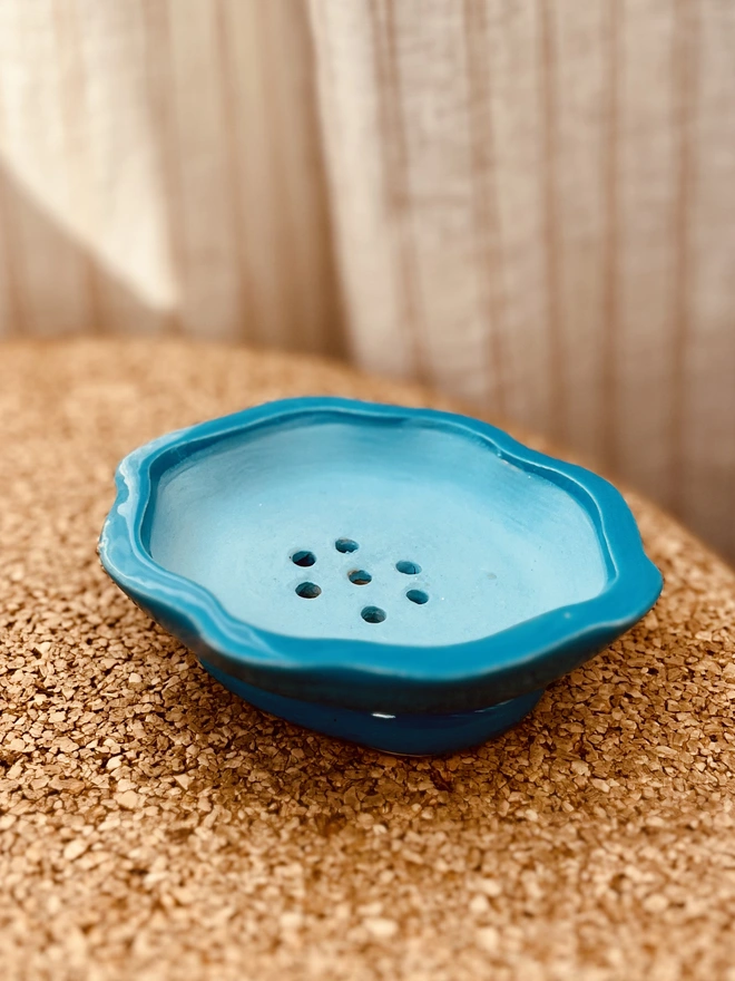 soap dish on a cork table with a fabric background. the soap dish is oval in shape with a wavy rim and has a bright blue outside and sky blue inside