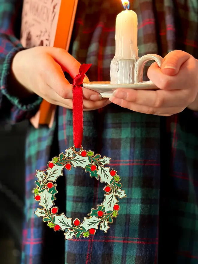 A child wearing a tartan dress holds a vintage-style enamel Christmas wreath ornament with green holly leaves and red berries, hanging from a red ribbon, alongside a small white candleholder with a lit candle.