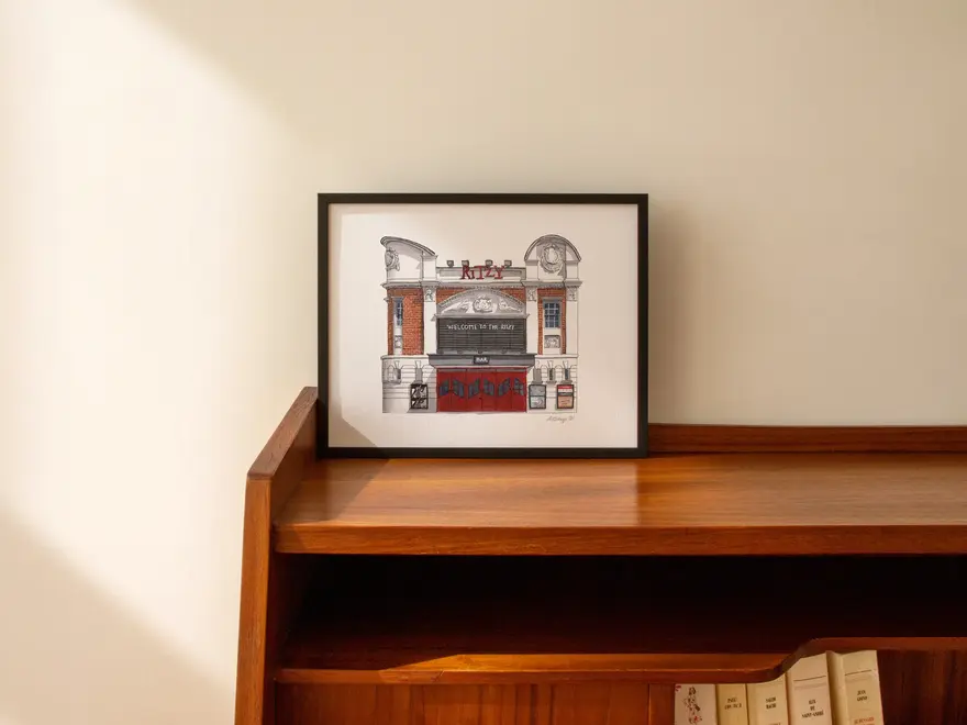 Brixton Ritzy Cinema Giclée Print (Unframed), a print in a black frame sitting on a wooden surface against a white backdrop. 