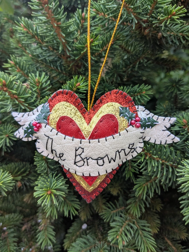 Hand stitched personalised heart Christmas decoration made from vegan-friendly faux leather, featuring a cream name banner, sparkly holly leaves, red berries and a gold hanging cord.