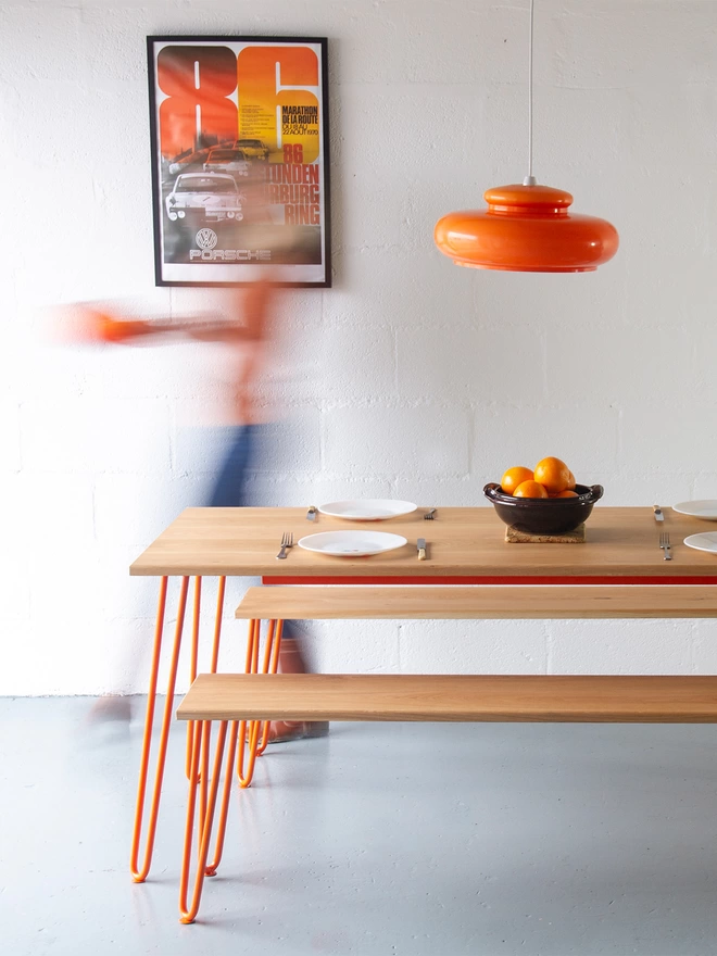woman walking away from a hairpin leg kitchen table with oak top and orange legs, with benches either side and an orange glass lamp shade above