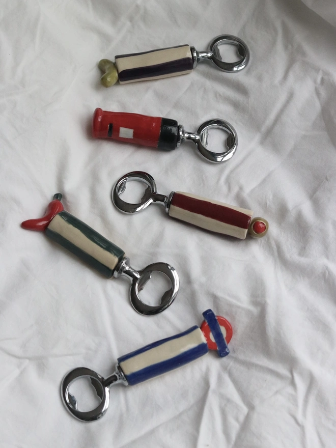 Tube, Olive, Chilli, Or Pickle Bottle Openers, a group of bottle openers on a white tablecloth. They each have a small food or fruit design. 