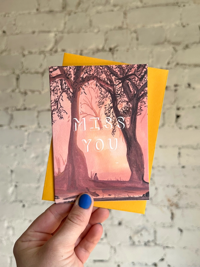 a greetings card featuring an illustration of a sunset through the trees with the text “miss you” with a deep yellow envelope