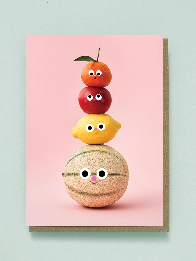 A totem of happy fruit on a pink background. A melon, lemon, apple & satsuma 