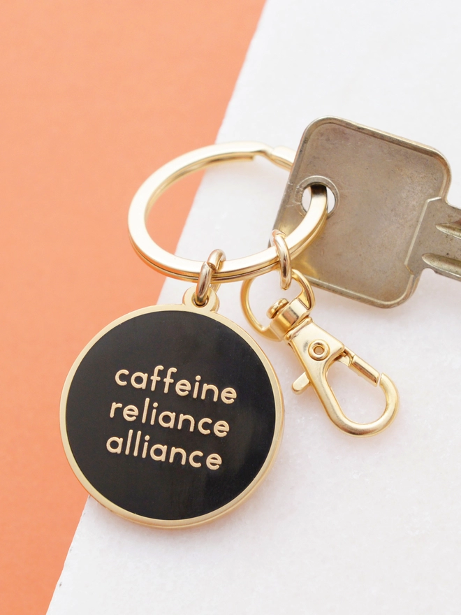 Caffeine reliance alliance enamel keyring laying on a marble and orange background