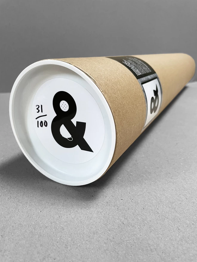 A cardboard tube laid on a plain grey background, containing the tea and biscuits ampersand with a sticker on the end depicting that.