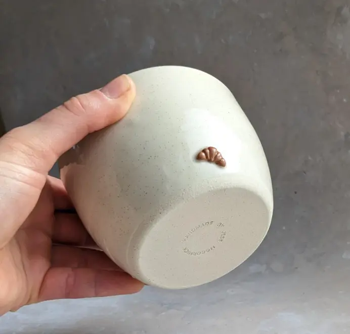 Vanilla bean–colored croissant coffee cup with a subtle croissant design, placed against a grey marble background.