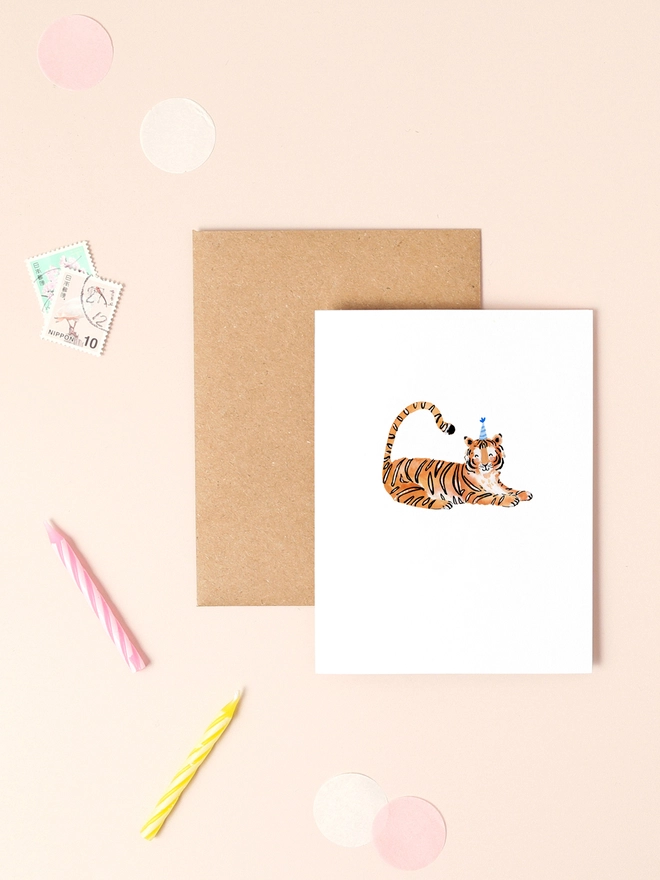 A playful illustration of a cheerful tiger lounging on its belly, wearing a festive blue party hat adorned with a pom-pom. The tiger's orange and black stripes are depicted in soft, flowing lines, adding to its friendly expression. The card's minimalist design features a neutral background, allowing the tiger and its party hat to take center stage.