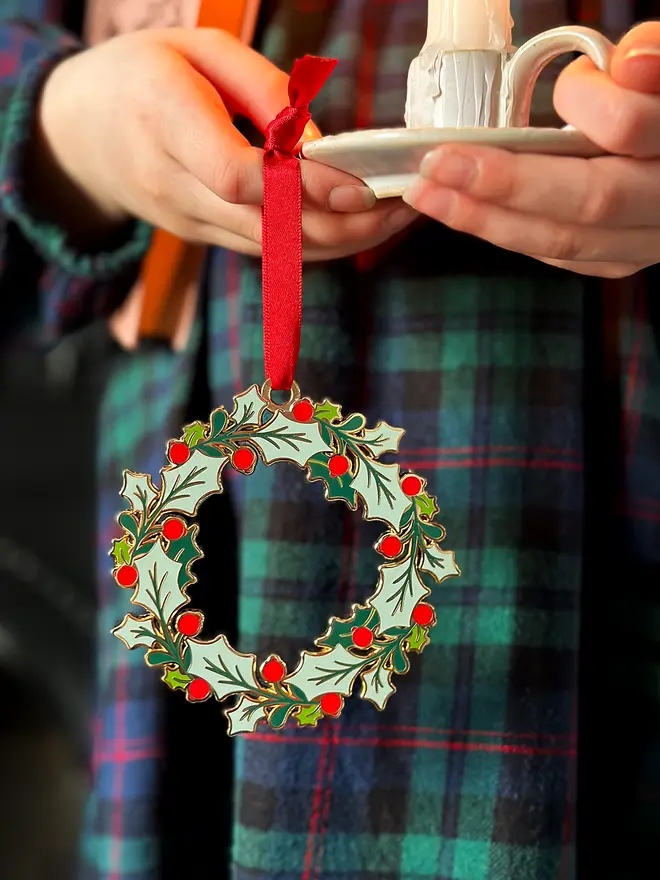 A child wearing a tartan dress holds a vintage-style enamel Christmas wreath ornament with green holly leaves and red berries, hanging from a red ribbon, alongside a small white candleholder with a lit candle.