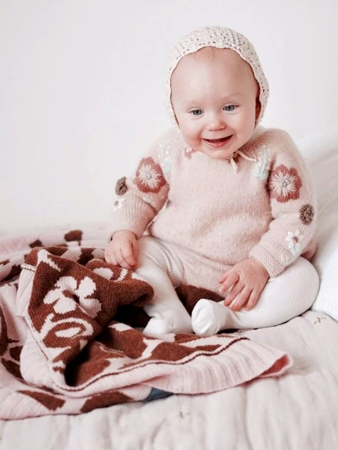 A smiling baby sits up on a bed, reaching out for the knitted blush briar rose baby blanket.