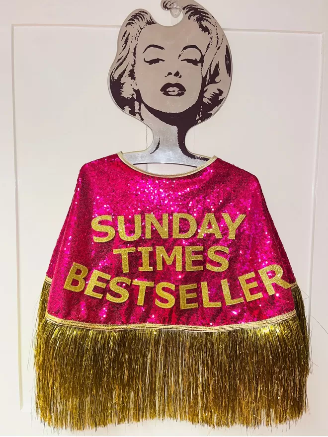 a midi cape with hot pink sequins, gold text reading 'SUNDAY TIMES BESTSELLER' and gold tinsel