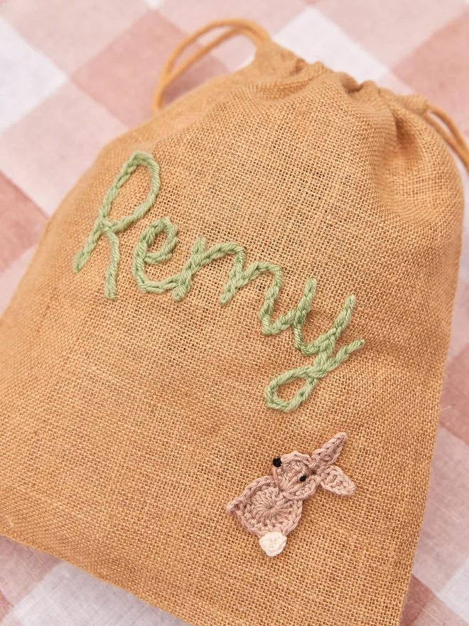 jute drawstring sack with brown knitted bunny decoration and green 'remy' name next to golden chocolate eggs on red gingham table cloth