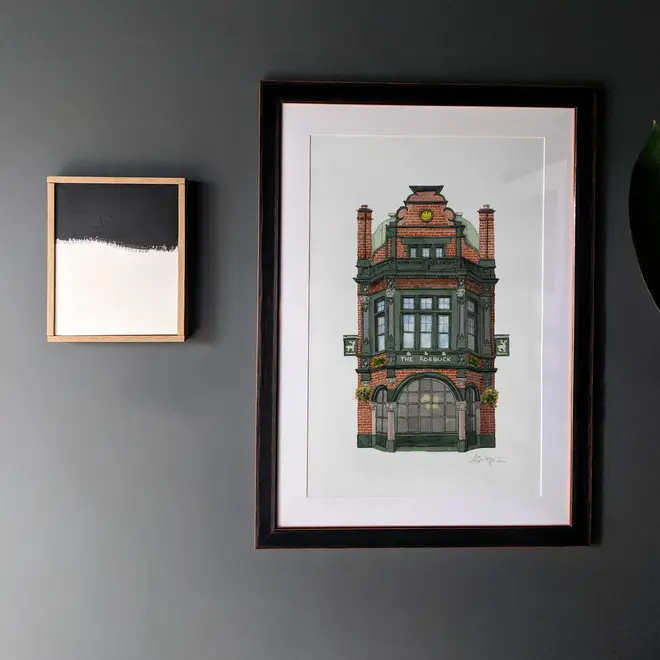 Borough The Roebuck Pub Giclée Print (Unframed), a print of a pub sitting in a black frame on a wooden surface against a black backdrop. It is accompanied by some another print. 