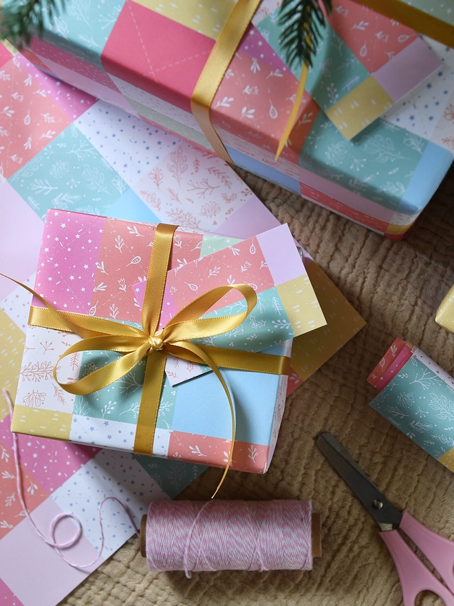 Gifts wrapped in gift wrap with a design of pastel patchwork quilt design is on a wooden table.