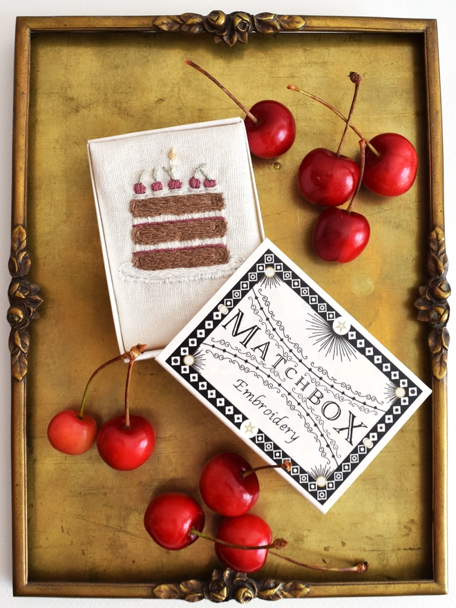 The finished embroidery placed inside the matchbox packaging for display.  Shown here on a vintage brass tray with scattered home grown cherries.