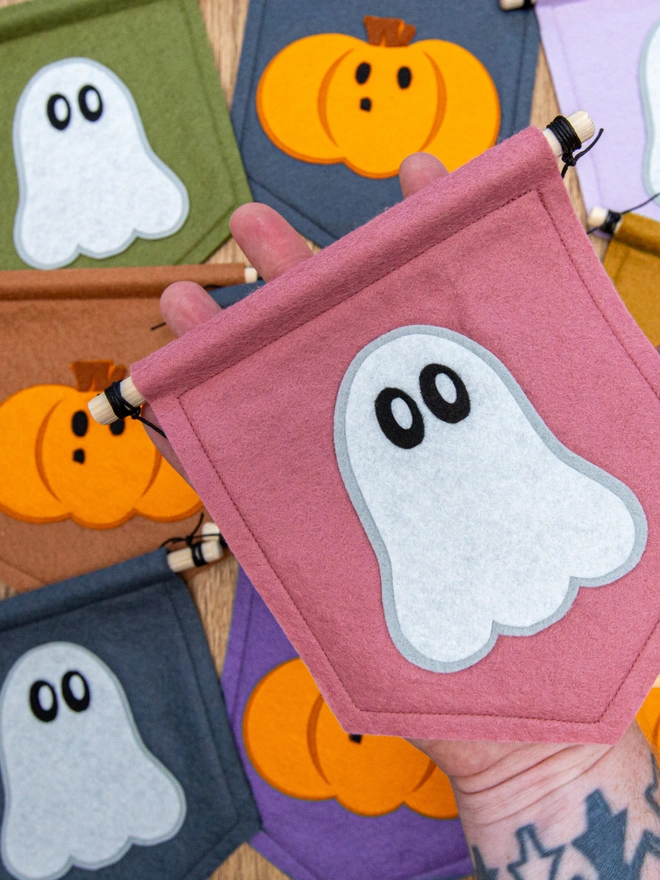 A cute looking halloween ghost with wide eyes on a rose pink banner. In the background are more banners in a range of other colours including mossy green and purple. There is also a cute pumpkin on some of the banners as an alternative.