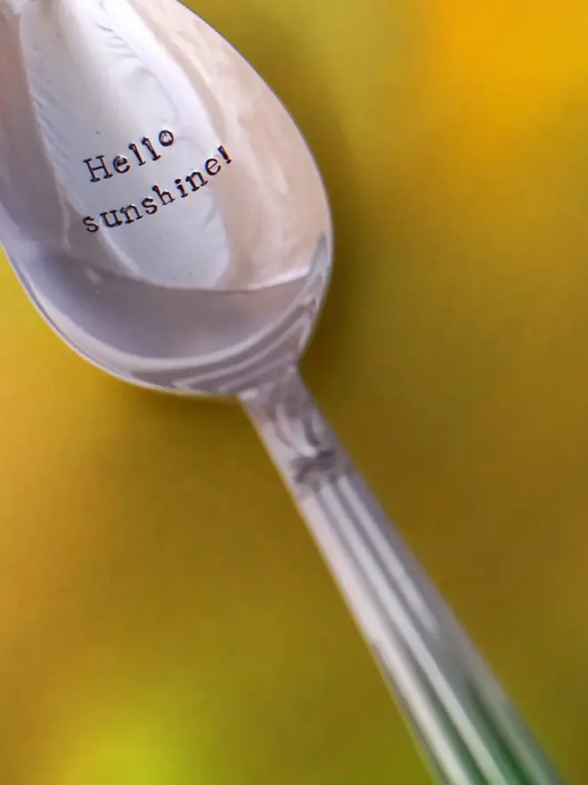 A vintage silver plated dessert spoon with the personalised hand stamped message reading Hello sunshine! The spoons has a decorative design on the handle and is on a yellow background.